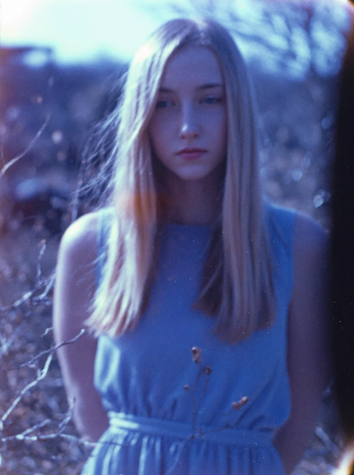 p0l2rd, Young woman, late 20s, fair skin, long, straight, blonde hair, partially obscured face, wearing a light blue, sleeveless dress, arms behind back, standing amidst thin, introspective mood, bare branches, outdoor natural setting, sunlight filtering through branches, overexposed and blurry photo with blue and purple tones, daytime