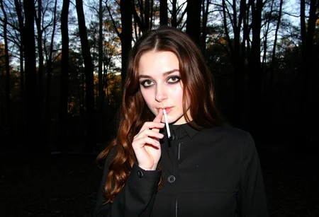 low-resolution photo,  shot on a mobile phone, noticeable graininess, no visible JPEG artifacts, some lens flare from the sunlight in the background. Young woman, early 20s, casual style, wearing dark trench coat, long wavy brown hair cascading over her shoulders, looking direct at the viewer, gothic heavy makeup, outdoors in the dark forest, autumn, holding a cigarette to her lips, smoking cigarette, sunlight filtering through the trees