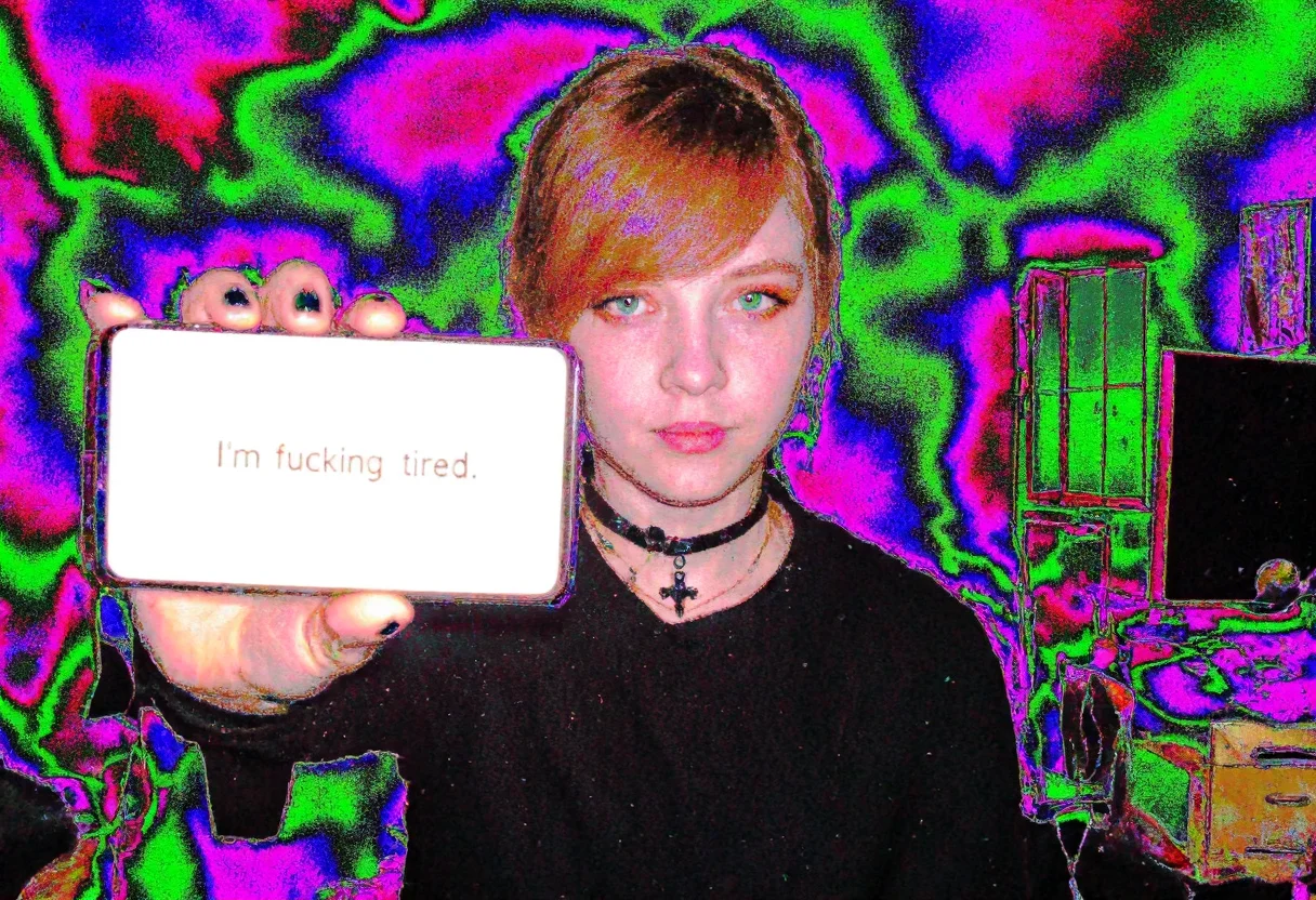 Circuit-bent/glitch art photograph showing a young woman, early 20s, easter european, fair skin, reddish-brown hair pulled back, side-swept bangs covering part of the forehead, dark eyeliner, orange eyeshadow, natural lip color, She also has a black choker and a black cross necklace. Her expression is neutral. room with furnitures and desk with computer and monitor and window. holding Samsung note10 with bright illuminated display to viewer, display showing text "I'm fucking tired". She has black nailpolish and rings on some fingers. artificial indoor heavy light, low light condition, underexposed face, face slightly shadowed, daytime. bright multicolored textures dominating the subject, wave-like color distortions, vivid neon color shifts with predominant green blue and magenta hues, distinct pixelation and edge artifacts, overall high digital corruption level