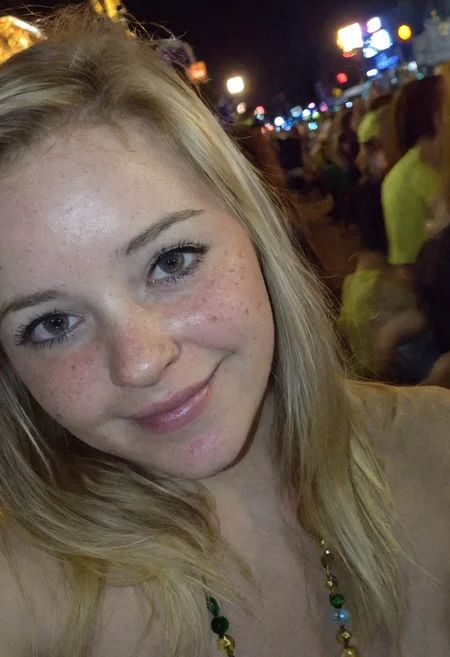 Amateur cellphone photography capturing a cute and attractive young woman with blonde hair at Mardi Gras during a stunning sunset. The scene is vibrant with the festive atmosphere of the parade, colorful beads, and lively crowds in the background. The woman has a playful smirk on her face, exuding confidence and charm, with subtle freckles (freckles:0.2) adding a touch of natural beauty. The photo is taken with a Samsung Galaxy in challenging lighting conditionsâpoor lighting, low light, and underexposedâresulting in a high-contrast image with deep shadows and bright highlights. The composition emphasizes the woman as the focal point, with the warm hues of the sunset casting a golden glow on her features, while the surrounding environment remains moody and atmospheric. The imperfections in the photo, such as graininess and slight blur, add to the raw, authentic feel of amateur photography.