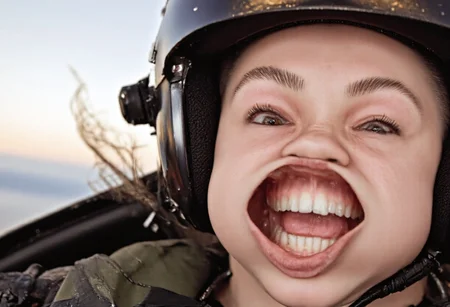 Photo of a female pilot inside a cockpit of a fighter jet flying at high speeds. She is wearing an RAF flight suit and helmet. She is a beautiful pale-skinned woman with freckles and brown eyes. w1nd8l0wn photo of a squinting woman's perfect teeth. windy. She is wearing clear lipgloss that accentuates her plump full lips. big wide open mouth. stretchy flappy lips fully exposing gums and teeth. realistic. high-definition. sunrise warm lighting. Surprised expression as she endures high-speed g-force.