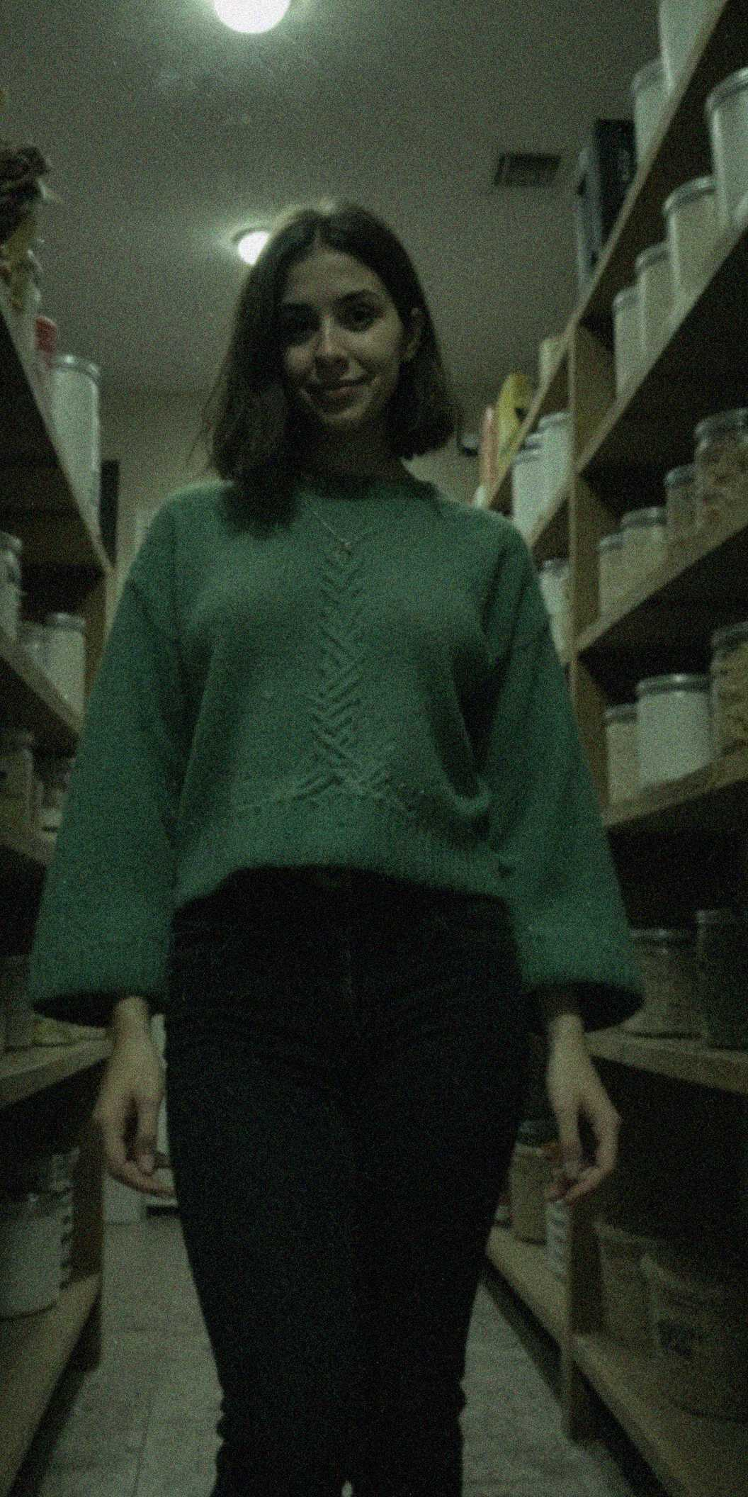 (raw, light leak, film grain, jpeg artifacts, low res, low quality:1.3), shot on GoPro Hero, , 20 year old woman, white woman, olive skin, thick body, bob hair, white hair color, forest green chunky knit oversized sweater, round neck, worn with black skinny jeans, walk-in pantry interior, floor-to-ceiling wooden shelves neatly organized with glass jars (flour, sugar, pasta), canned goods stacked, baskets holding root vegetables, hanging bunches of dried herbs, cool dim lighting, Midnight