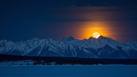 A breathtaking photograph of a full moon rising over a snow-covered mountain range, casting an ethereal glow over the landscape. The sky is deep and dark, creating a striking contrast with the bright orange moon, tinged with subtle hints of golden yellow.
The towering mountains, blanketed in pristine snow, stand majestically, their rugged peaks illuminated by the soft, ambient light. Scattered trees add depth and texture to the composition, their silhouettes accentuating the vastness of the winter terrain.
In the background, the setting sun casts a warm, golden glow, blending seamlessly with the moonlit sky. The interplay of cool blue shadows and soft, warm highlights creates a visually stunning contrast, evoking a sense of tranquility and awe.
The overall mood is peaceful and serene, with the stillness of the snowy wilderness enhanced by the luminescent beauty of the moon and the fading light of the sun. A masterfully composed, high-resolution cinematic landscape, capturing the grandeur of nature's quiet majesty.