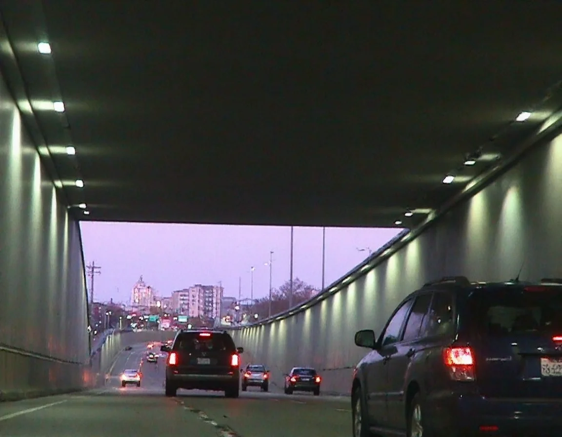 m8vic2, The image depicts a tunnel. The tunnel leads towards New York city in the background, lit up in the evening light with rows of car lights visible. Multiple vehicles are captured driving through the tunnel, including a dark-colored car in the foreground on the right, a black SUV in the middle ground, and several other vehicles further in the background on a multi-lane road. Inside the tunnel, light strips run along the ceiling, casting an even glow. The scene portrays an urban setting, indicative of evening commute in a metropolitan area.,