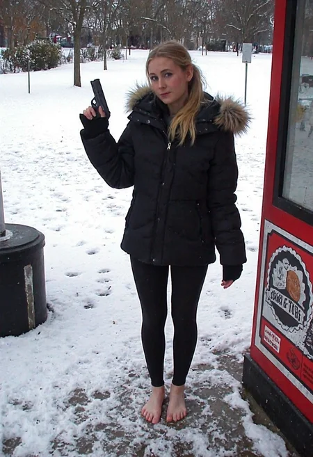 m8vic2, Young woman, early 20s, casual winter attire, wearing a black puffer jacket with a faux fur hood, tight black pvc leggings, no shoes, barefoot on wet snowy ground. She stands at a food stall outdoors, holding a handgun right hand, left hand gesturing at viewer. Blonde long ponytail, natural makeup. Background shows a park, snow-covered ground, footprints, overcast weather, trees in distance, wintery atmosphere.,