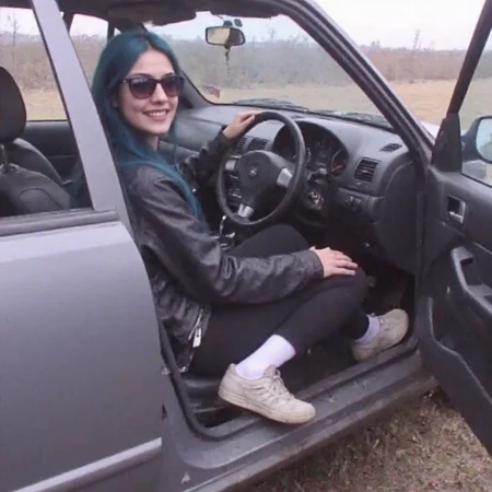 v8s, woman in her early 20s sitting inside a car in passenger seat, open car door, hand on knee, jacket, sunglasses, blue shoulder-length hair, pants, socks, sneakers, light smile, very happy pose, dutch angle, cowboy shot. Amateur quality, Analog distortion, low resolution, Chromatic aberration, tape noise, Grainy texture, Frame jitter, muted colors, overexposed foreground