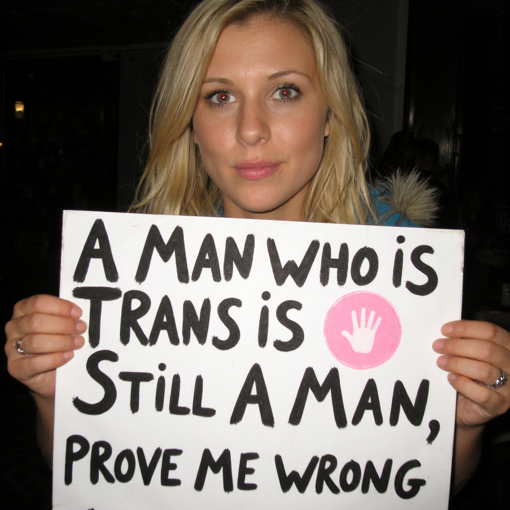 Amateur Party Photography of a woman with blonde hair and red eyes from camera flash looking intimidating, holding a sign in bad handwriting with the text: ((A Man Who is Trans Is Still A Man, Prove Me Wrong)).