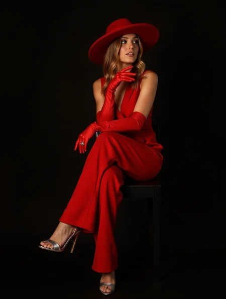 A woman poses elegantly on a black chair, wearing a vibrant red outfit consisting of a sleeveless jumpsuit, long red gloves, and a wide-brimmed red hat. Her long, wavy hair falls over her shoulders, and she looks slightly to the side with a confident expression. She wears silver high-heeled sandals, adding a touch of glamour to her bold aesthetic. The background is dark, highlighting the vibrant red of her outfit.