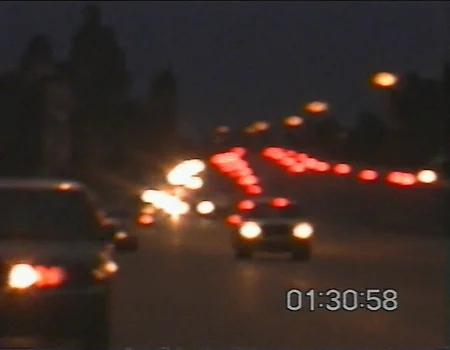 v8s, blurry nighttime cityscape, defocused urban lights, red and amber bokeh, vhs aesthetic, digital timestamp reading 01:30:58 in bottom right corner, horizontal scan lines across light sources, low resolution quality, chrominance shift creating color bleeding, dark atmospheric mood, grainy texture, nighttime traffic lights, vintage video recording, slight combing artifacts, nostalgic lo-fi ambiance, deep shadows, muted color palette