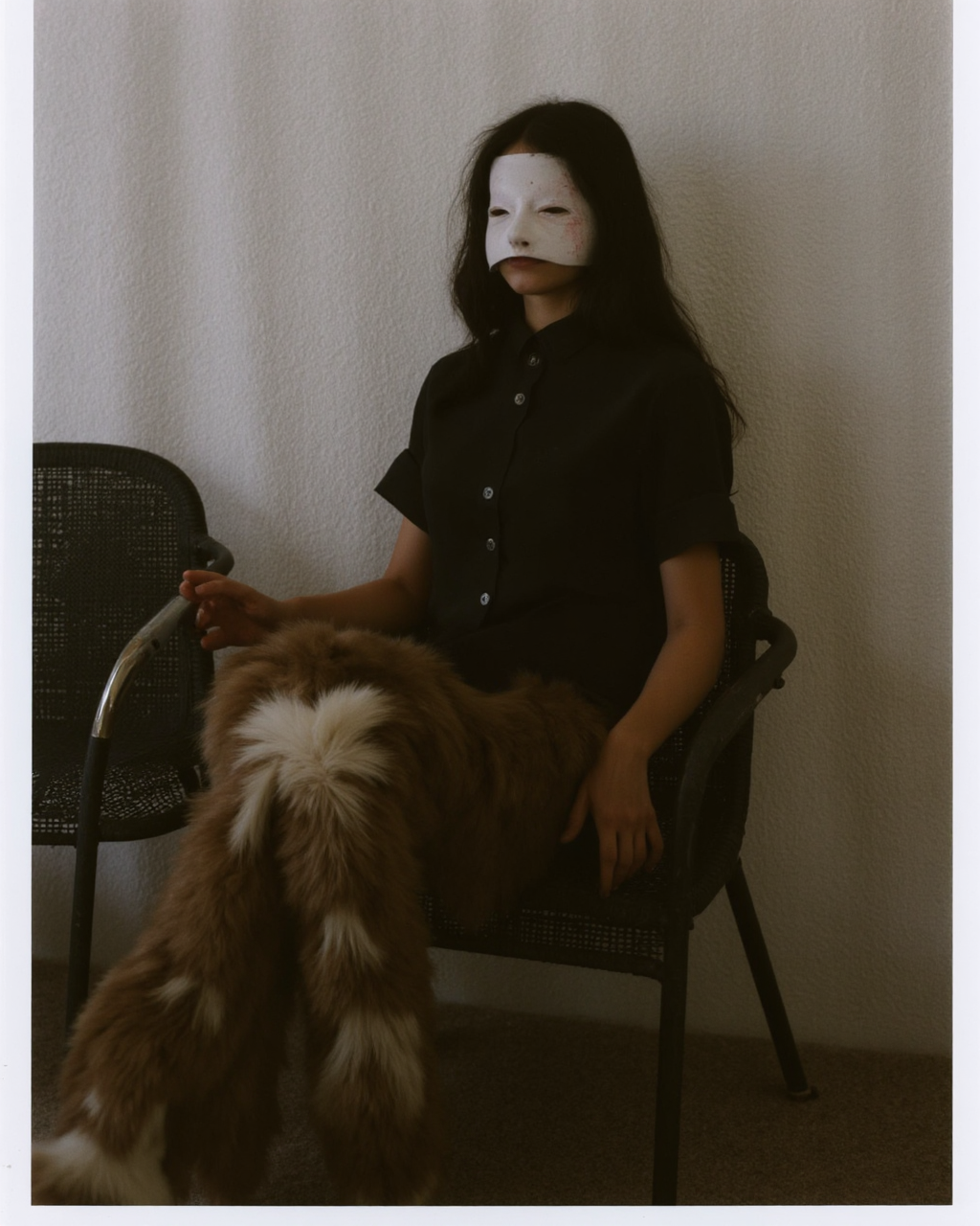 Kaos, The image is a photograph with a surreal, eerie atmosphere. The subject is a person, likely a young woman, with a face mask that obscures their features, giving them an unsettling, almost inhuman appearance. They have long, dark hair cascading down their shoulders. The person is dressed in a dark, short-sleeved, button-up shirt, which contrasts with the lighter, ethereal background. The individual is seated in a dark, wicker chair with a mesh backrest. Their legs, covered in fur, are positioned in a relaxed, slightly spread stance, with one foot resting on a chair. The fur appears to be a mix of brown and white, adding to the otherworldly feel of the image.