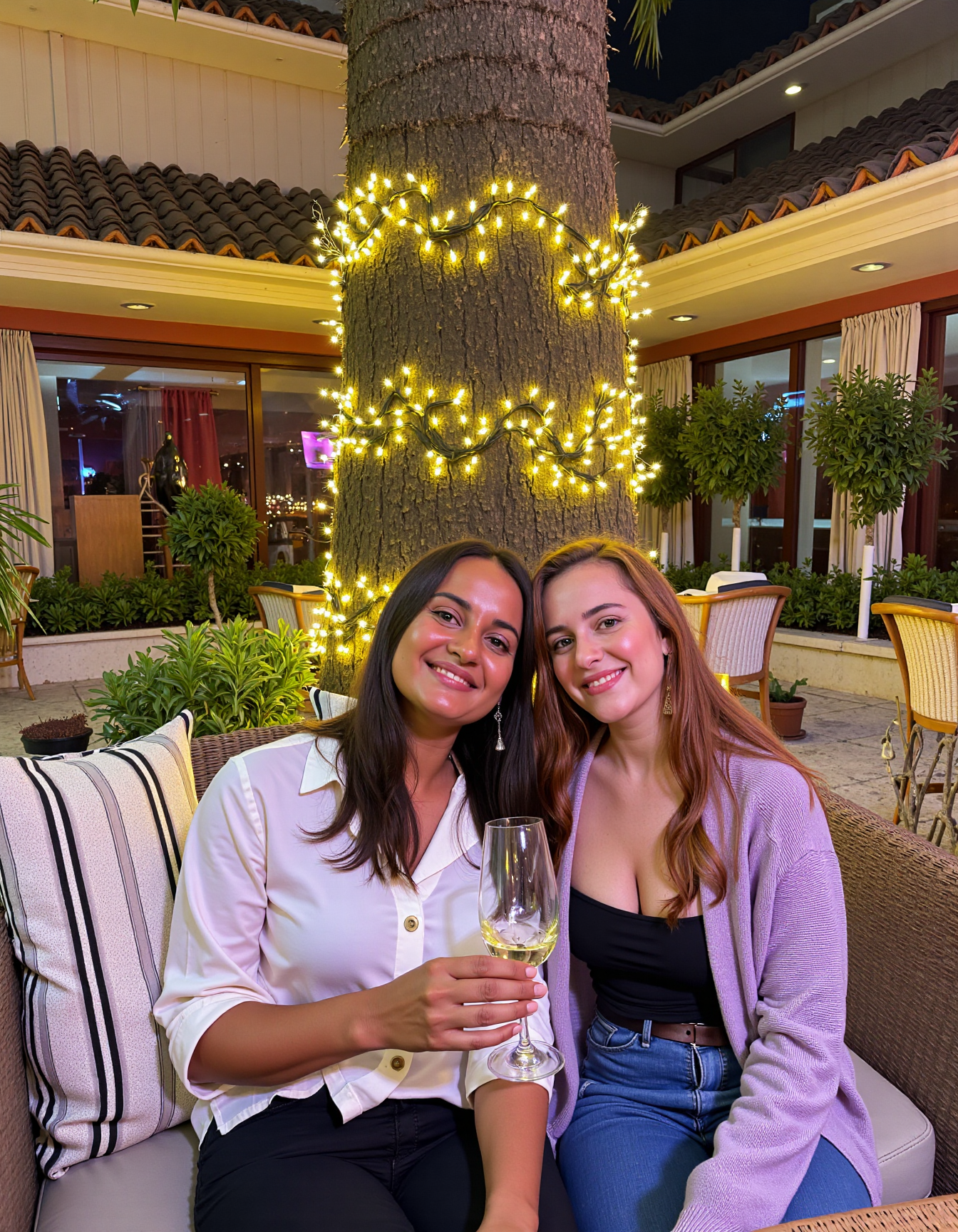 A medium shot photo of two women sitting outdoors at a cozy, warmly lit patio in the evening, with soft, glowing lights wrapped around a tree behind them creating a charming atmosphere. The image is rich in detail, with warm tones and clear textures throughout. The woman on the left has a medium build with tan skin, long dark brown hair falling loosely over her shoulders, and is smiling slightly while holding a glass of white wine near her lips. She is wearing a light, silky button-up shirt paired with dark pants. The woman on the right has fair skin, a slim build, and long reddish-blonde hair, also worn loose. She is smiling warmly at the camera, holding a glass of red wine in her right hand, and wears a soft-textured lavender cardigan over a black top. Both women are seated closely on a striped cushioned bench, with their posture relaxed and comfortable. The background features a large tree with sparkling string lights wrapped around its trunk and branches, adding a magical touch to the scene. The setting suggests a private outdoor seating area, with elements like terracotta-tiled roofing, greenery, and rattan chairs contributing to a serene, inviting vibe. The overall composition is balanced, with the women framed against the softly glowing tree, and the lighting enhances the warm, intimate feel of a quiet evening shared among friends. The details are vivid, with textures of clothing, skin, and background elements clearly visible, while the warm lighting creates a relaxed and joyful mood <lora:amateurphoto-v5-14-15-1-1:0.4>