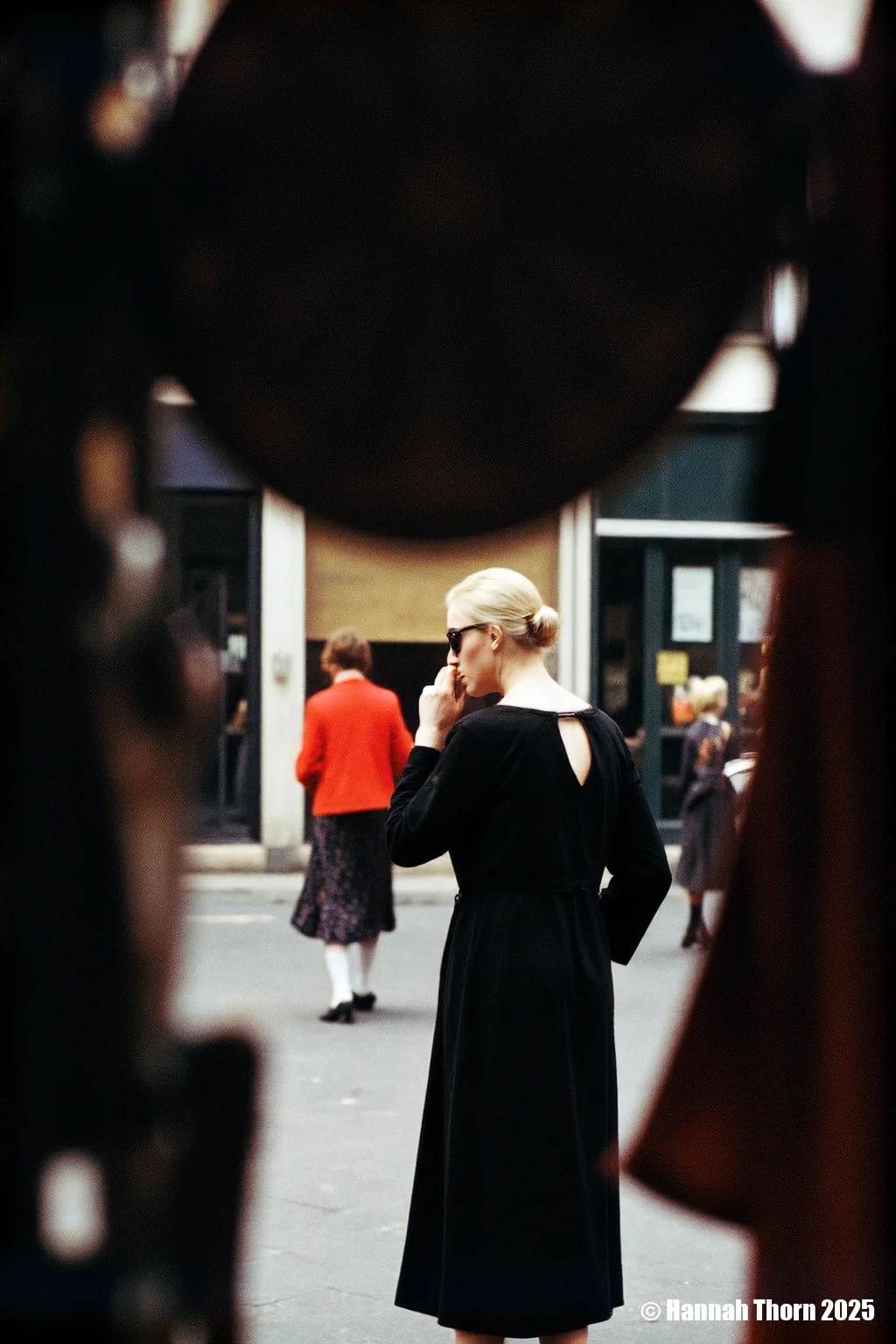 The picture is a street photograph, likely a candid shot, capturing a moment in a public space. A woman, seen from the back and slightly to the side, is the central figure. She wears a simple, dark, possibly black, long-sleeved top or dress with a low v-back. Her blonde hair is pulled back, and she's wearing dark sunglasses. Her right arm is bent, and her hand is raised to her face, either touching her cheek or adjusting her glasses. In the background, another woman, smaller and slightly out of focus, walks away from the camera. She's wearing a vibrant red jacket and a dark, patterned skirt that falls below the knee. Her legs are visible, and she appears to be wearing light-colored stockings and dark shoes. The overall style of the photograph is reminiscent of street photography from the mid-20th century. Several elements contribute to this impression: * **The use of color:** The colors are somewhat muted, suggestive of older film stock. The pop of red from the woman in the background contrasts with the darker tones of the central figure and the overall scene. * **The focus and depth of field:** The central woman is in sharper focus, while the background and the second woman are softer, creating a sense of depth and isolating the main subject. * **The framing and composition:** The photograph is cropped in a way that emphasizes the central woman while also including the other woman in the background as a secondary element of interest. The dark, undefined shapes in the foreground suggest the photographer may have been shooting from behind a partially obscured vantage point, like through a window or foliage, adding a layer of mystery and candidness. * **The grainy texture:** A noticeable grain or noise is present throughout the image, adding to the vintage feel and again hinting at the use of film. * **The moment captured:** The scene is everyday and unposed, characteristic of street photography's aim to capture fleeting moments of real life. The woman's gesture suggests a natural, unselfconscious action, rather than a posed portrait. The photograph evokes a sense of mystery and intrigue, partly due to the obscured foreground, the candid nature of the shot, and the fact that the central woman's face is partially hidden by her hand and sunglasses. The image invites the viewer to speculate about the context of the scene and the women's thoughts or actions. The style is observational and documentary, focusing on the interplay of light, shadow, and human presence in a public environment. It has a timeless quality that speaks to a particular era of street photography, perhaps the 1950s or 1960s. <lora:_Flux\Saul_Leiter_Streetphotography.safetensors:1.0:1.0>