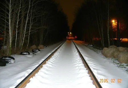 Snow-covered train tracks, barren landscape, leafless trees on both sides, scattered debris including tires to the left, darkness surrounds with sparse lighting from distant streetlights, wintry night, orange timestamp 2005/02/02 in the lower right corner, cold atmosphere, amateur quality, slight blur in the foreground.,