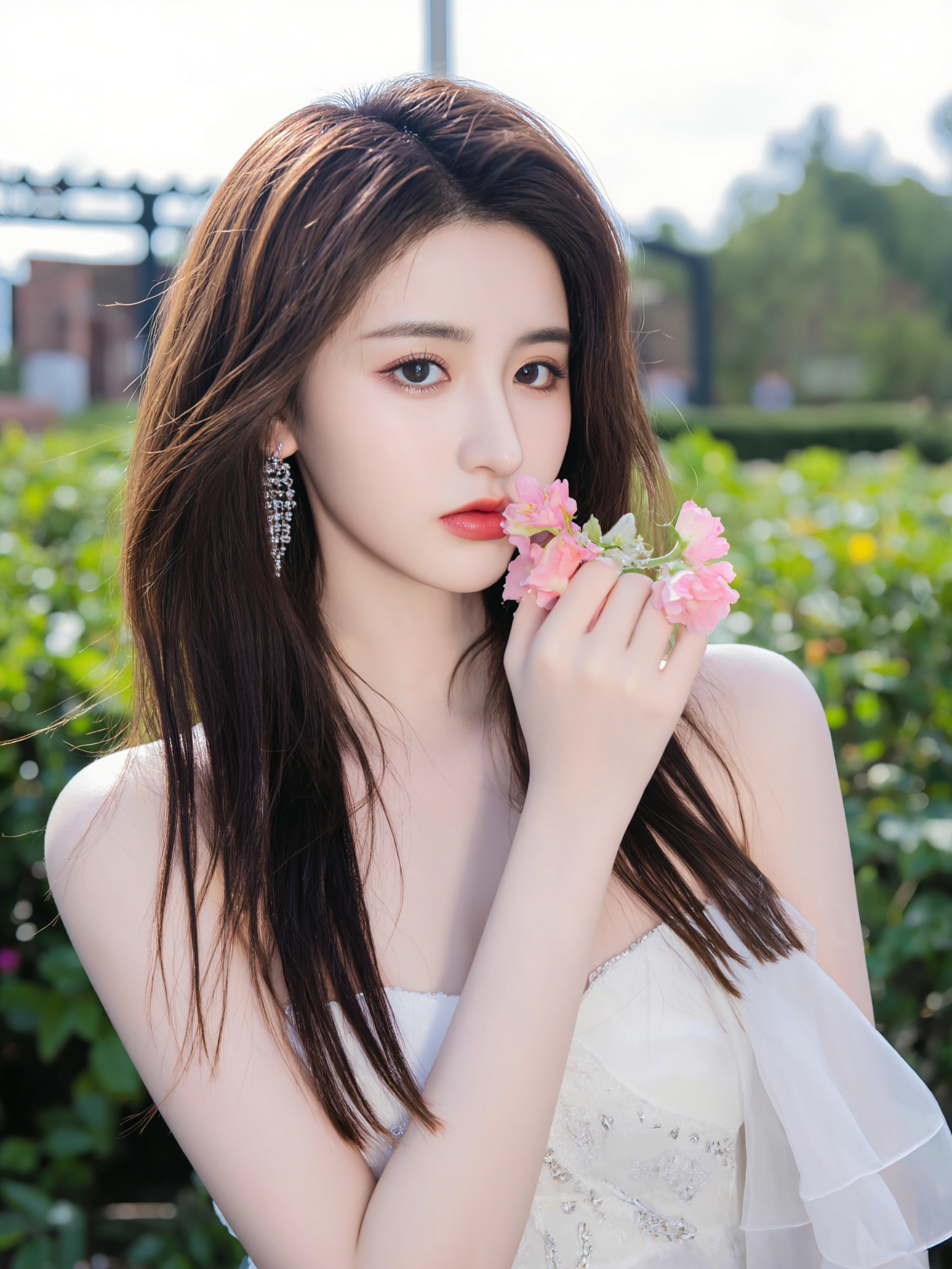 This is a high-resolution photograph featuring a young Asian woman with fair skin and long, straight, dark brown hair. She is wearing a white, off-the-shoulder dress with a sweetheart neckline, adorned with delicate silver embroidery. The dress accentuates her slender figure and small breasts. She is holding a pink flower close to her face with her right hand, while her left hand rests gracefully by her side. Her makeup is subtle, with a focus on her striking red lips, adding a pop of color to her otherwise neutral look. She wears large, sparkling earrings that catch the light.

The background is softly blurred, showcasing a garden setting with greenery and a hint of a circular structure, possibly a gazebo or a pergola, in the distance. The lighting is natural, likely from the sun, creating a soft, warm glow on her skin and highlighting her delicate features. The camera angle is slightly low, looking up at her, which adds a sense of intimacy and elegance to the composition. The image employs the rule of thirds, with the woman's face positioned in the upper third of the frame, drawing the viewer's attention to her serene expression and the flower she holds.