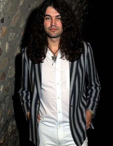 <lora:IAN_GILLAN:1>, This is a photograph of a man with long, dark, wavy hair that falls past his shoulders. He is dressed in a white, long-sleeved shirt and a black and white striped blazer. The blazer has vertical stripes that alternate between black and white. He wears a black tie with a silver pendant in the shape of a butterfly, which hangs prominently from his neck. The man has a serious expression and is slightly smiling. His left hand is casually placed in his pants pocket, while his right arm hangs loosely by his side.
The background of the photograph is a dimly lit, textured wall that appears to be made of stone or brick, with a slightly rough texture. The lighting in the image is soft, casting gentle shadows that enhance the texture of the man's hair and the striped pattern of his blazer. The overall color palette is muted, with the dominant colors being black, white, and shades of grey. The photograph has a candid, natural feel, capturing a moment of relaxed introspection. The image's composition is centered on the man, with the background being slightly blurred, focusing attention on him.