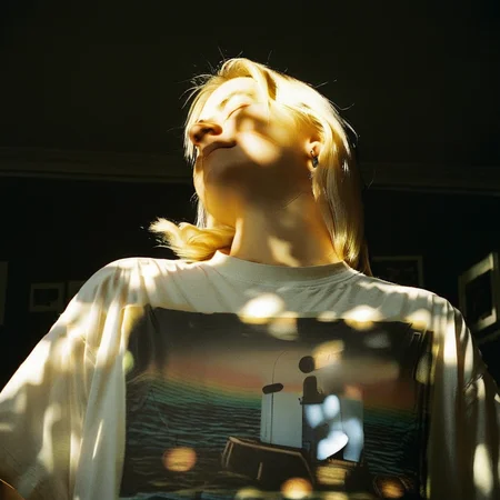 FLMFX, Film photo, Analog photography, 35mm film, Grainy texture, Film grain, Soft lighting, Vintage look, Washed colors, Light leaks, Natural shadows, 1girl, solo, long hair, shirt, blonde hair, closed mouth, jewelry, white shirt, upper body, closed eyes, earrings, indoors, medium hair, shadow, sunlight, t-shirt, realistic, light, dark, print shirt, photo (object), sidelighting, photorealistic