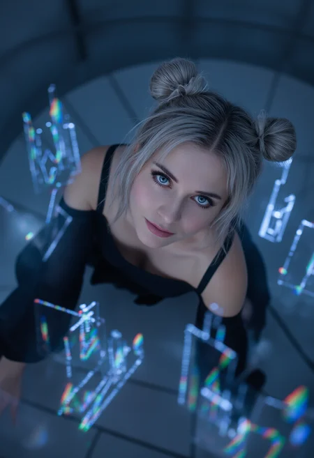 Glamour photography featuring a young hungarian woman, in a brutalist interior setting with holographic lighting highlighting her slim features at an angle from the top. The eye-level shot's focal point is the woman's face, giving only a glimpse of her black cold-shoulder top. Her round light blue eyes are accentuated by dark mascara complemented by long false lashes in a goth style, alongside black lipstick on her full lips. Her silver hair is styled into a big twin-buns style, with side-swept bangs. Her expression is intrigued and bold, her head slightly turned down, gazing directly at the viewer, with a flirty smile adding to the alluring mood of the image. Set on a spiraling brutalist stairwell with glitching digital candles in a wireframe design.