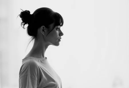 an aesthetic, minimalist depiction of a female profile in side view. she wears a t-shirt. The focus is on the soft contours and calm, monochromatic tones. The scene feels mystical and dreamy, almost as if viewed through a delicate mist, with gentle light accentuating the silhouette. The background is diffuse and creamy white, drawing attention to the elegant simplicity of the figure.