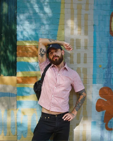 A man with a beard and tattoos standing in front of a colorful striped wall, posing confidently with one hand on his hip and the other resting on his thigh. he is wearing a red and white striped button-up shirt, black pants, and a black bag slung over his shoulder. his body is slim, and he has a beard. he has several tattoos on his arms, including one on his left arm and another on his right arm. he also has a cap on his head and a ring on his finger. the man is looking directly at the camera with a serious expression. the background is a vibrant outdoor setting with a striped wall in shades of blue, green, and yellow.
ðï¸ [Inpainted with Loras]: flux1-depth-dev-lora
ð¯<lora:flux\loratools\flux1-depth-dev-lora.safetensors:0.75:0.75>