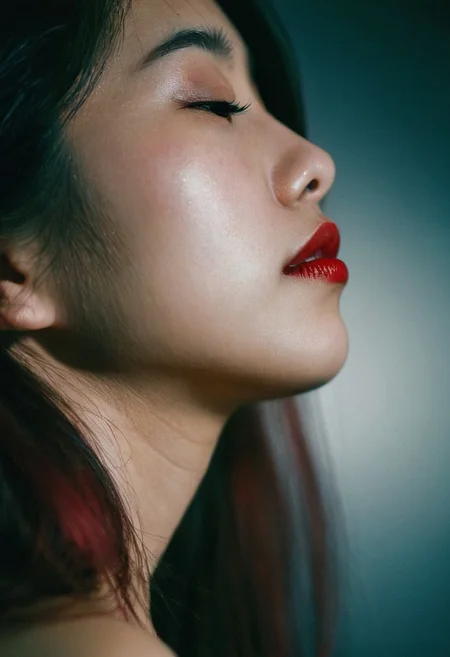 shot on fujifilm x100vi, medium close-up portrait, 28 years old asian girl, side view, sharp jawline, pale glowing skin, deep red lipstick, soft highlights, soft natural lighting, dim dark-ish background, Superia Xtra 400 Film, film emulation, 35mm film, grainy, halation, Bloom, high dynamic range, , film photography, 23mm focal length, iso 100, f/2.8, (sharp details, high quality, 4k, intricate details, highly-detailed)