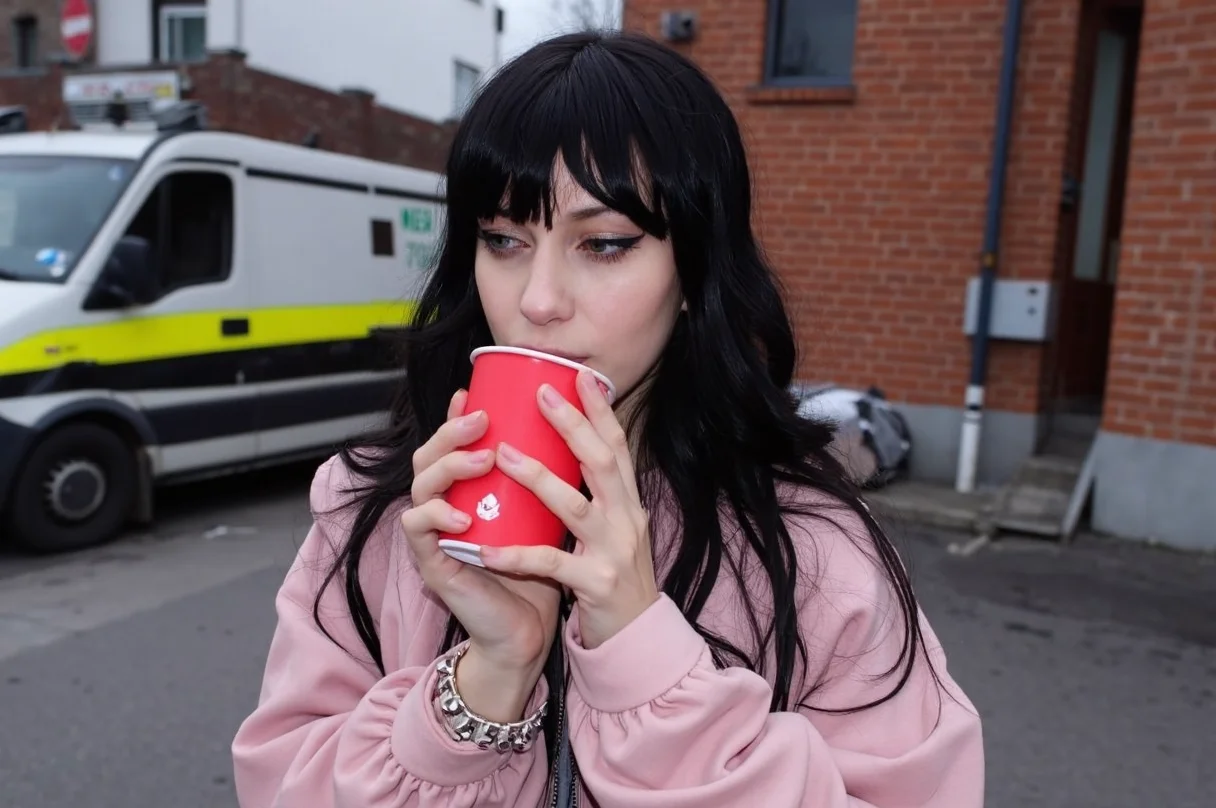 Young woman, mid-20s, pale complexion, long dark hair with bangs, wearing a vintage oversized pink bomber jacket, holding a red cup close to her lips, adorned with silver studded bracelets, subtle makeup with pronounced eyeliner, slightly parted lips, standing, outdoor urban backdrop, parked van and brick building in background, overcast lighting, blurred, casual, candid, amateur quality, smeared details, in motion