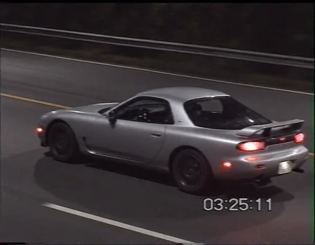v8s, rear three-quarter view of a silver sports car mazda rx7. The car appears to be driving on high speed on a dimly lit highway at night, with a broken taillight emitting an orange glow. A black grooved rear wing and aftermarket wheels suggest the car is modified, likely belonging to someone in the drifting or tuner car . The interior is slightly illuminated, showing racing seats with a roll cage. The background reveals a guardrail and a dark, empty road, intensifying the isolation of the scene, low lighting, in motion, smeared details.  horizontal tearing and segmented scanlines all over still image of vhs footage, time base error distortion, analog video artifacting, low resolution and dark lighting, white text "03:25:11" in bottom right corner