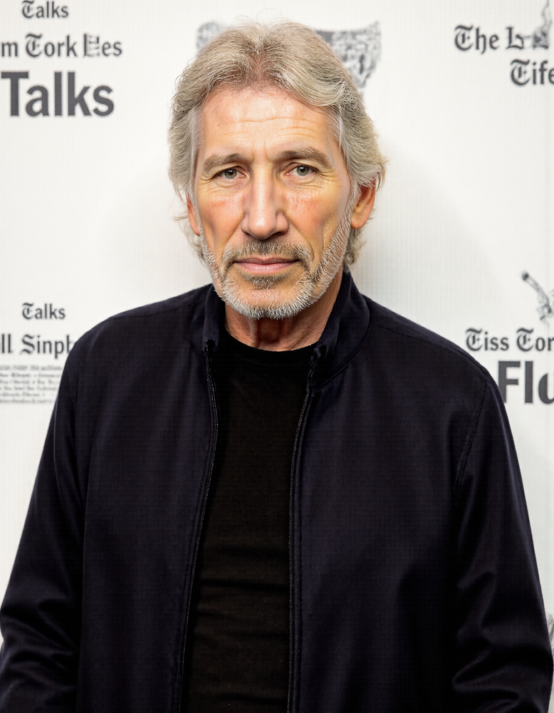 <lora:ROGER_WATERS:1>, This is a color photograph of an older man with white hair and a full, white beard, standing in front of a white backdrop adorned with repeated text. The text reads "The New York Times" and "Talks, " indicating a promotional or event-related setting. The man is wearing a dark-colored, zip-up jacket over a black shirt, which contrasts with his white hair and beard. His expression is neutral, and he appears to be looking directly at the camera. The lighting is even, highlighting his facial features and the textures of his clothing. The background is simple and clean, emphasizing the subject. The image captures a candid moment, likely taken at a talk or interview session, as suggested by the backdrop. The overall composition is straightforward, focusing on the subject with minimal distractions, allowing the viewer to focus on the man and his presence.