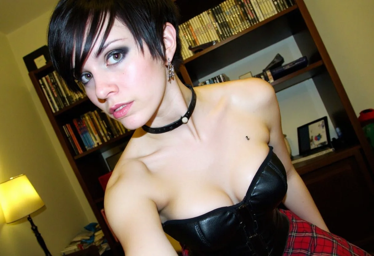 low-resolution photograph taken in a cluttered, possibly bedroom setting, showing a young woman with short, dark hair. She appears to be of Caucasian ethnicity with a light skin tone. The woman is dressed in a black leather corset that accentuates her small to medium-sized breasts, and a red plaid skirt, she is slightly leaned forward and reveal her cleavage. She has a black leather choker with a metal ring around her neck, and a silver ring in her left ear. Her lips are slightly parted, showing a small silver stud in her lower lip.
In the background, there are shelves filled with various items, including what looks like books and DVDs, giving the room a somewhat disorganized appearance. The lighting is dim, casting a yellowish hue over the scene, possibly due to a lamp or light source in the room. The overall ambiance suggests a rebellious or alternative style. The image is slightly blurred, possibly due to poor lighting or a shaky camera