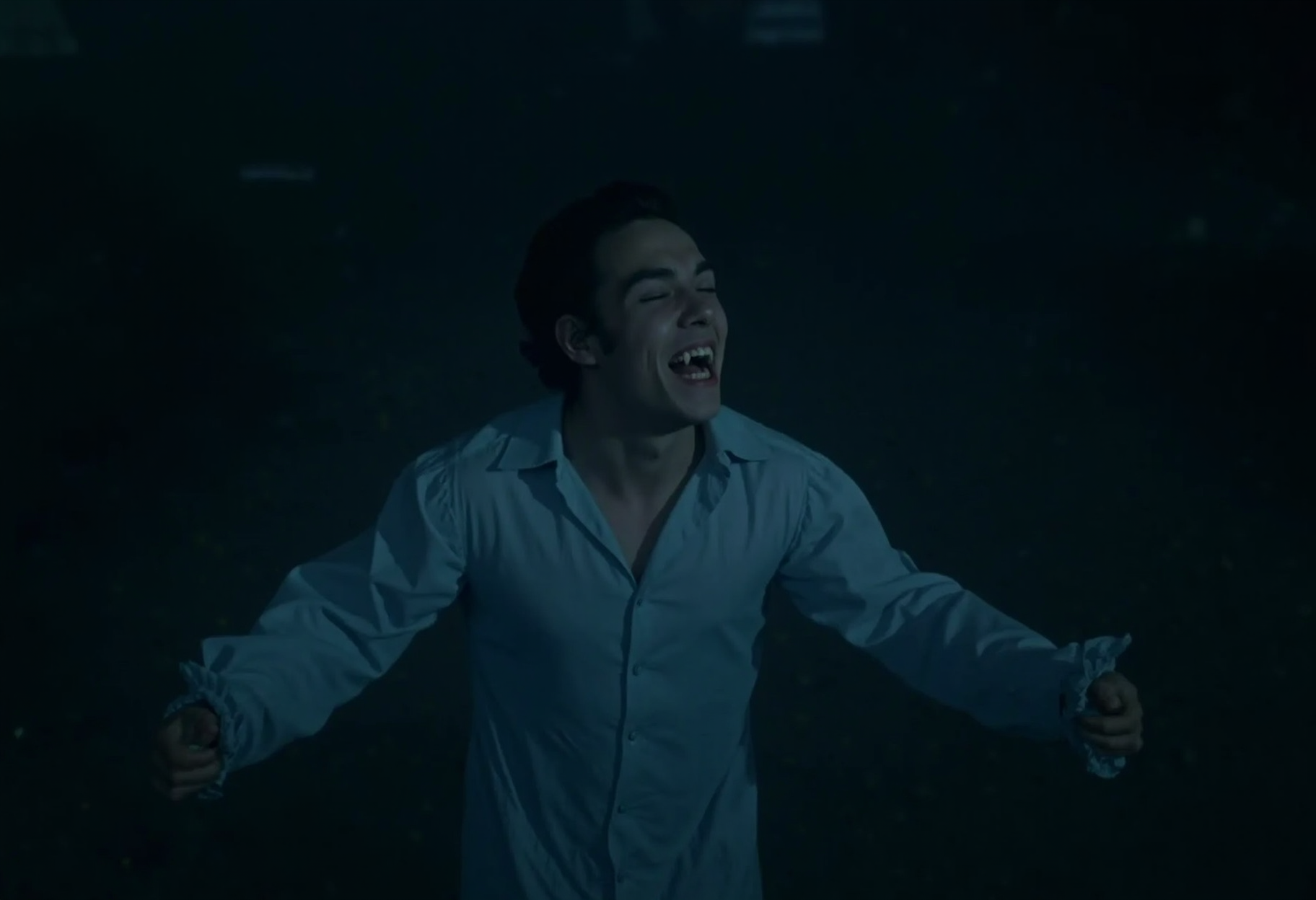medium shot of a young vampire man standing on a cemetary in the rain at night, wearing a victorian nightshirt, arms spread, looking up with eyes closed, smiling showing fangs, pale skin, taken from above. low light at night <lora:hammer-000073:1>