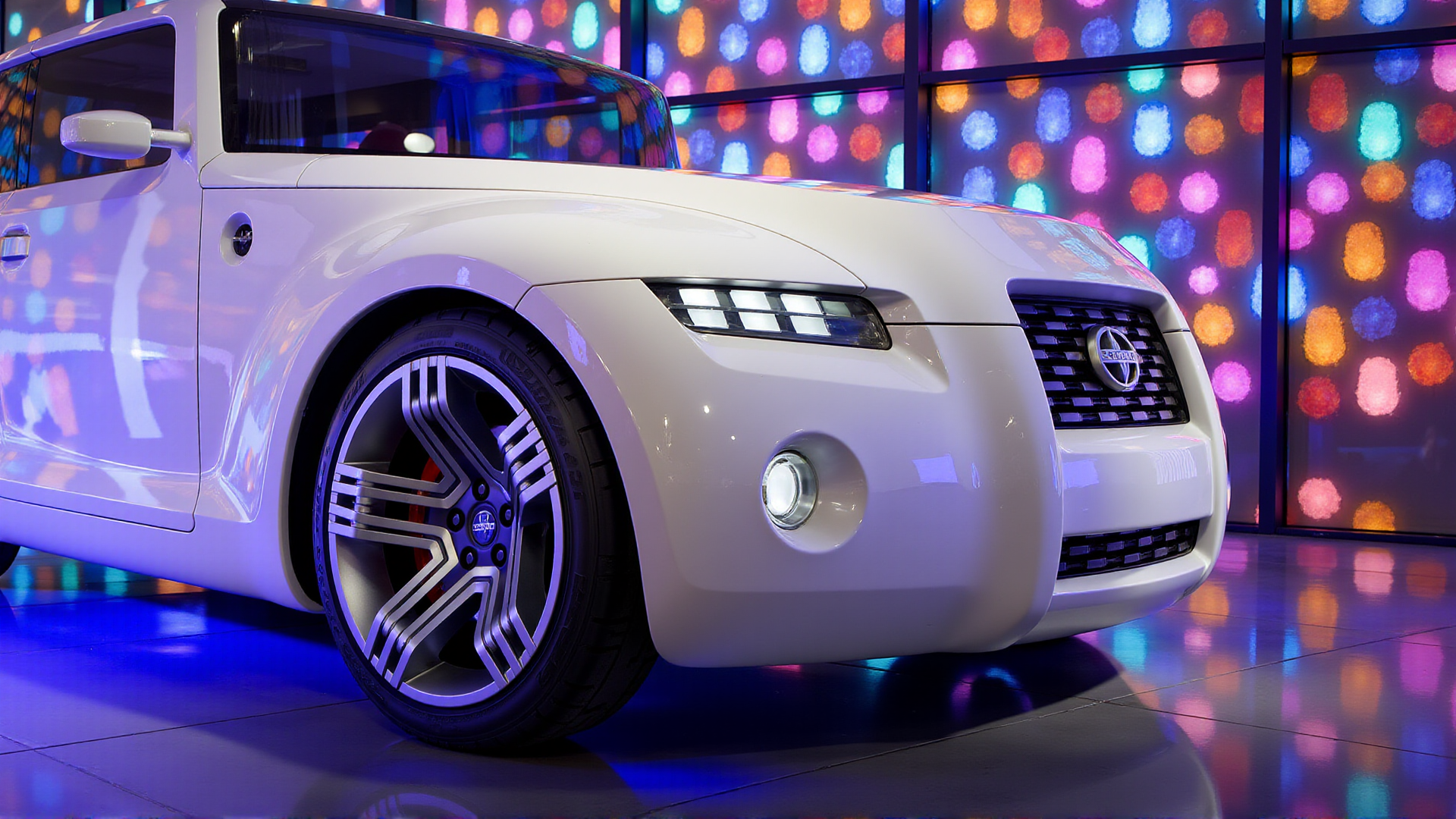 A dazzling close-up of the Scion Hako Coupe Concept in a studio filled with prismatic crystal structures. The carâs livery is a reflective diamond white, refracting the rainbow-like light bursts from surrounding crystals. The shot focuses on the narrow LED headlights and the intricate geometric grille, with colorful light rays dancing across the surfaces. The polished multi-spoke alloy wheels and glowing fog lights add sharpness to this vibrant and luxurious setting
 <lora:ral-hkcpcncpt_000004000:1>