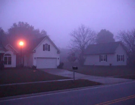m8vic2, Residential neighborhood at dusk, foggy evening setting, two houses visible, left house larger, white exterior, arched window, dark roof, right house smaller, grey exterior, black shutters, streetlamp glowing orange on the left side, bare trees silhouetted in the background, dark mailbox, empty driveway, dim lighting conditions, purple-blue haze dominates, outdoor urban environment, low visibility, street and grass barely discernible, overcast sky., 