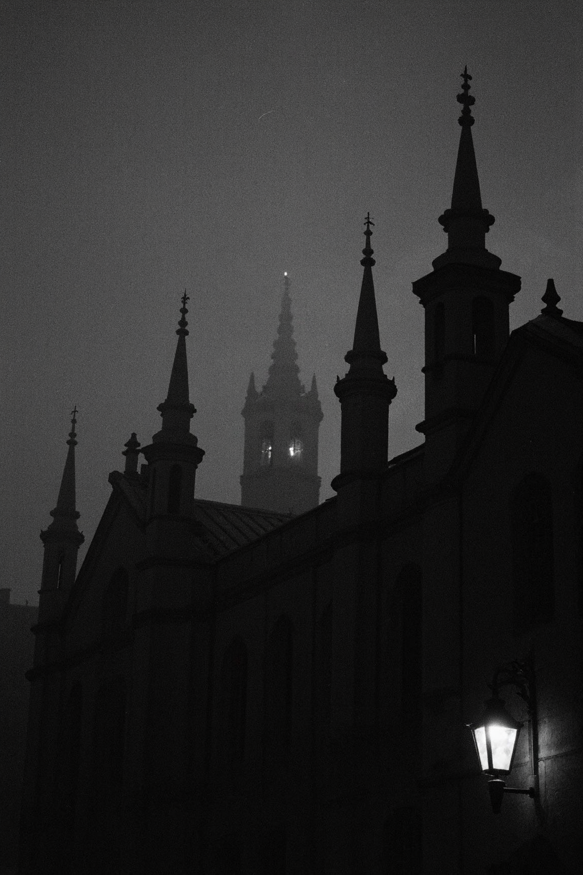 8n8log,Dark silhouettes of gothic architecture towers with pointed spires and ornamental finials against a gloomy night sky, single illuminated lantern or light fixture visible on right tower creating soft glow, multiple tower structures of varying heights visible, medieval or castle-like building features, dramatic moody atmosphere, low visibility conditions, possibly fog or mist present, black and white or monochrome photography, underexposed image creating high contrast between structures and sky, architectural details barely discernible in the darkness, nighttime setting, atmospheric gothic ambience, cathedral towers. film photography aesthetic, film grain effect prominent throughout image, high contrast lighting creating dramatic shadows, grainy film-like texture, professional photography technique, dramatic chiaroscuro lighting effect