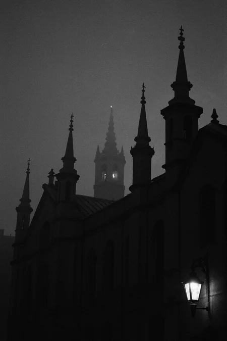 8n8log,Dark silhouettes of gothic architecture towers with pointed spires and ornamental finials against a gloomy night sky, single illuminated lantern or light fixture visible on right tower creating soft glow, multiple tower structures of varying heights visible, medieval or castle-like building features, dramatic moody atmosphere, low visibility conditions, possibly fog or mist present, black and white or monochrome photography, underexposed image creating high contrast between structures and sky, architectural details barely discernible in the darkness, nighttime setting, atmospheric gothic ambience, cathedral towers. film photography aesthetic, film grain effect prominent throughout image, high contrast lighting creating dramatic shadows, grainy film-like texture, professional photography technique, dramatic chiaroscuro lighting effect
