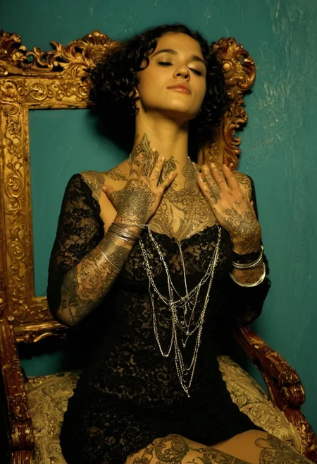 The image is a photograph of a young woman with a medium-light skin tone and curly, dark hair styled in a short bob. She is seated on an ornate, upholstered chair with intricate wooden carvings and a dark green fabric. She wears a sheer, black, lace dress with intricate patterns, revealing her figure underneath. The dress has long, flowing sleeves and a deep neckline that accentuates her décolletage.
Her tattoos, visible on her arms and hands, are intricate and detailed, featuring floral and possibly abstract designs. She has a series of silver bangles on her wrists, and around her neck, a large, ornate necklace with several layers of silver charms dangles.
She is sitting with her hands gently touching her temples, a contemplative expression on her face, looking down. The background features a teal-colored wall with a rough, textured finish, giving an artistic, slightly industrial vibe. Above her, on the wall, hangs a vintage, sepia-toned portrait of a woman in an ornate, golden frame, contributing to the vintage, artistic ambiance.
The lighting is soft yet dramatic, casting a warm glow that enhances the richness of the colors and textures in the scene.