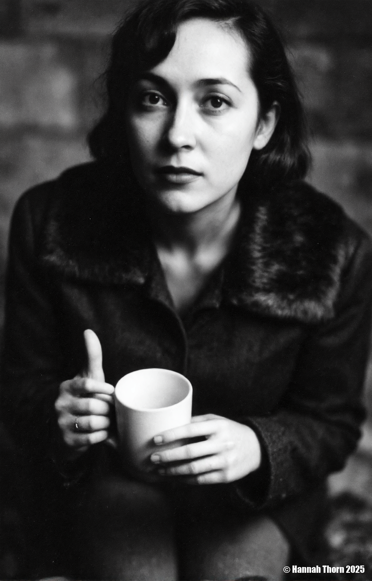 A black and white photo from a portrait camera angle about a woman holding a cup, looking directly at the viewer with a serious expression. the woman, who appears to be in her late 20s, has long, wavy hair and is wearing a black coat with a fur collar. she is sitting in the middle of the image, with her upper body visible, and her hands are holding a white mug. the background is blurred, with a dark, moody atmosphere, and the focus is on the woman's face and the mug she is holding. the image has a minimalist aesthetic, with no other elements present in the scene.<lora:_Flux\Saul_Leiter_BW.safetensors:1.0:1.0>