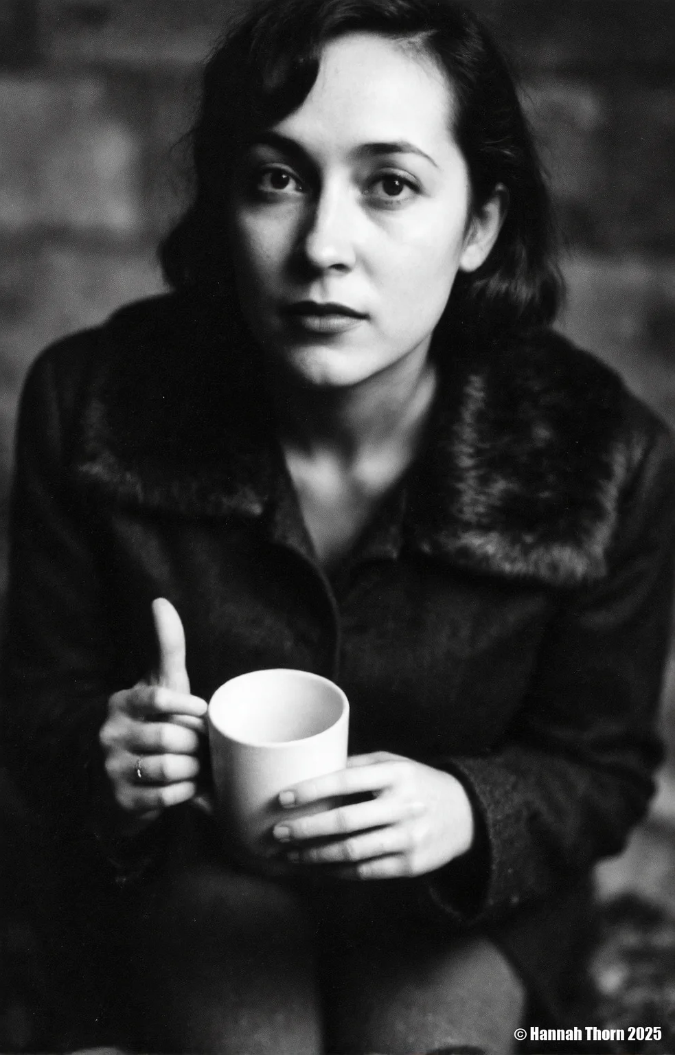 A black and white photo from a portrait camera angle about a woman holding a cup, looking directly at the viewer with a serious expression. the woman, who appears to be in her late 20s, has long, wavy hair and is wearing a black coat with a fur collar. she is sitting in the middle of the image, with her upper body visible, and her hands are holding a white mug. the background is blurred, with a dark, moody atmosphere, and the focus is on the woman's face and the mug she is holding. the image has a minimalist aesthetic, with no other elements present in the scene.<lora:_Flux\Saul_Leiter_BW.safetensors:1.0:1.0>