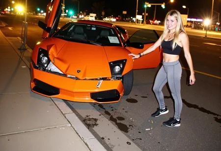 medium-resolution photo, shot on a mobile phone, slightly grainy, with minor visible JPEG artifacts and noise in the darker areas. The image captures an orange sports car lamborghini murcielago heavily damaged in the front, parked askew on a curb at a city intersection. Various angular elements of the damaged hood, express the severity of the impact. The car's right door is open, and there are noticeable wheel marks, debris, and liquid on the road, likely from the impact. Surrounding environment includes a paved sidewalk in the foreground and blurry, partially illuminated street with traffic lights and moving vehicles in the background, street lights casting a yellowish glare, suggest night time in an urban setting, dynamics of urban accidents, car culture. on foreground in front of the car is standing young woman and smile, dynamic pose, she is blonde with long hair, wear grey leggings and black crop top and sneakers