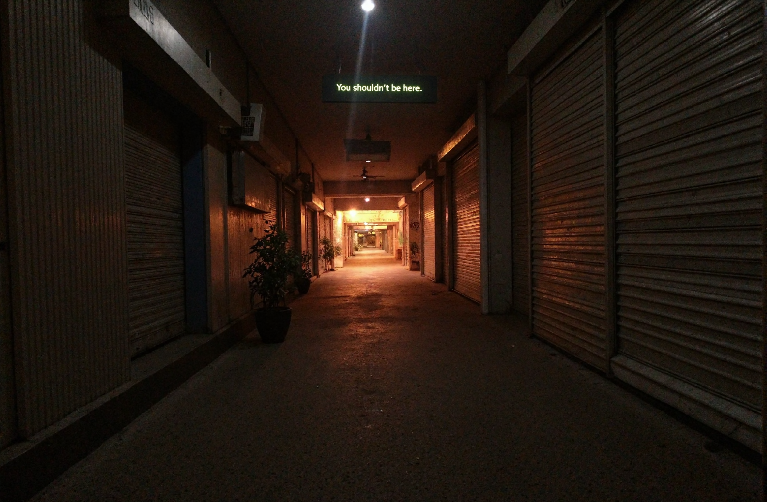 l3n0v0. wide-angle 18 mm interior shot, deserted shopping-mall corridor after closing time, rows of closed metal roller shutters, “You shouldn't be here” signage half-lit, lonely potted plant for scale, moody tungsten floodlights casting deep orange glow and long shadows, pockets of near-black darkness at the edges, soft volumetric beam slicing through dust motes, early-2000s CCD sensor vibe with slight noise and bloom, low-key cinematic lighting, mirror’s-edge-meets-y2k minimalism, gritty urban isolation mood. amateur quality, candid style