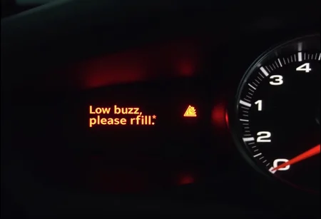 pov of a driver loocking at the car dashboard with the gasoline gauge on reserve with warning light on and an message saying "Low buzz, please refill", at the night with low light. The focus is on the gasoline gauge. The message is in red color and is a warning.