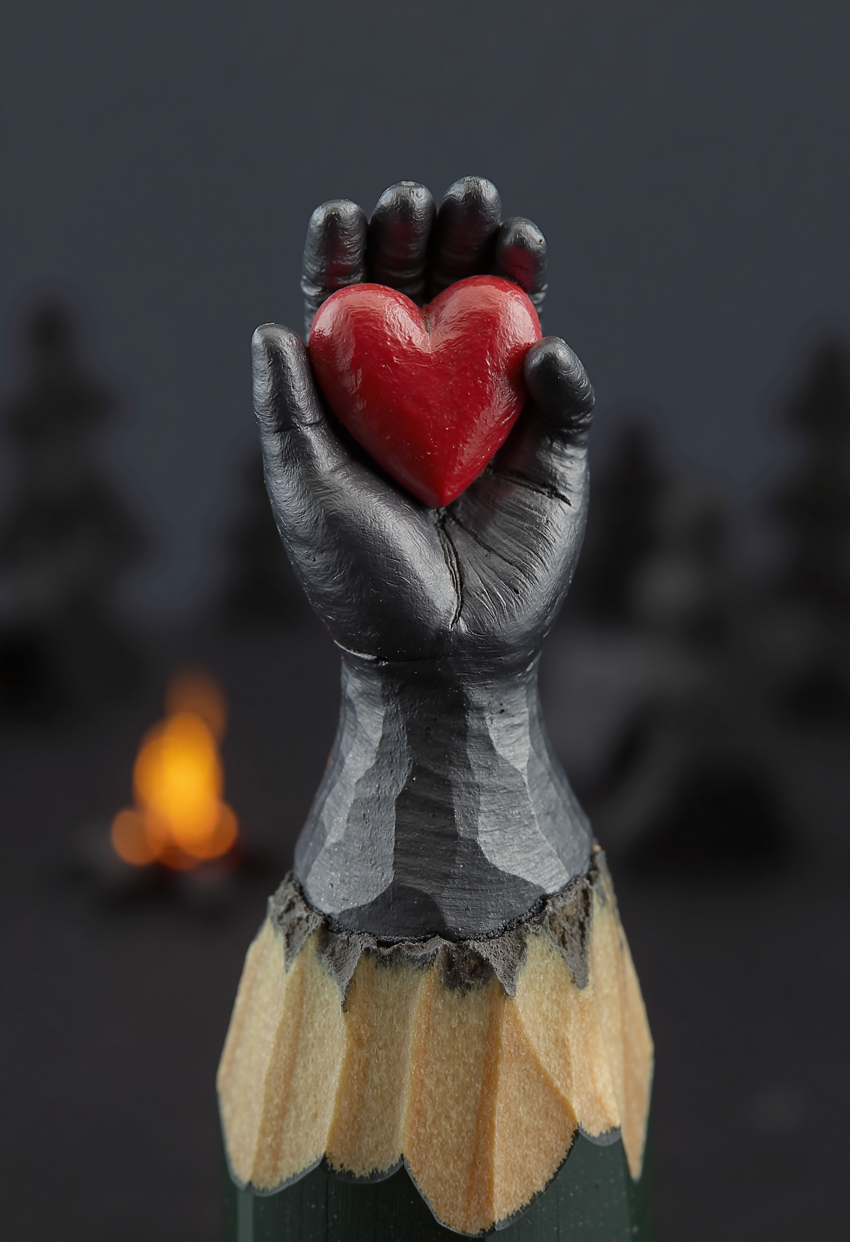 Mini sculpture on the tip of the pencil, a gray hand holding a heart in its palm. The heart is painted red. The background is dark gray, you can faintly see a campfire and a dark forest. 