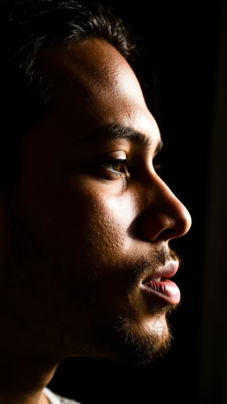 An ultra-realistic photograph of a manâs face in profile, his expression calm and reflective. His skin has a deep, bronze tone with subtle imperfections such as faint acne scars and a slightly uneven texture. His short beard is meticulously groomed, and the individual hairs are clearly defined. The light from a nearby lamp casts a soft glow on his cheekbone, while the shadow on the other side of his face creates depth and dimension, enhancing the lifelike quality of the image. , aidmarealisticskin