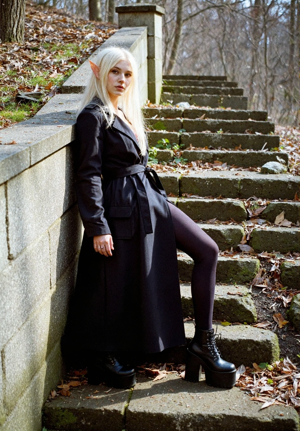 8n8log, Elf-like young woman in loose trench black coat, long straight platinum blonde hair, long pointed ears, dark leggings, demonia platform black boots, seductive facial expression, standing in extravagant pose, leaning against stone mossy wall, body angled sideways, her gaze directed to the viewer, outdoor forest setting, sunlight casting shadows, stone mossy destroyed steps in foreground, rocky ground, bare trees, natural lighting, amateur quality, dutch angle. film photography aesthetic, film grain effect prominent throughout image