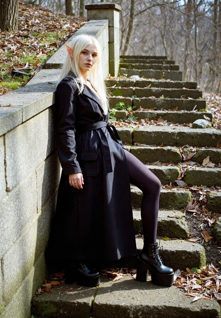 8n8log, Elf-like young woman in loose trench black coat, long straight platinum blonde hair, long pointed ears, dark leggings, demonia platform black boots, seductive facial expression, standing in extravagant pose, leaning against stone mossy wall, body angled sideways, her gaze directed to the viewer, outdoor forest setting, sunlight casting shadows, stone mossy destroyed steps in foreground, rocky ground, bare trees, natural lighting, amateur quality, dutch angle. film photography aesthetic, film grain effect prominent throughout image