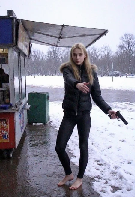 v8s,  Young woman, early 20s, casual winter attire, wearing a black puffer jacket with a faux fur hood, tight black pvc leggings, no shoes, barefoot on wet snowy ground. She stands at a food stall outdoors, holding a handgun right hand, left hand gesturing at viewer. Blonde long ponytail, natural makeup. Background shows a park, snow-covered ground, footprints, overcast weather, trees in distance, wintery atmosphere.. Amateur quality, Analog distortion, low resolution, Chromatic aberration, tape noise, Grainy texture, Frame jitter, muted colors, overexposed foreground