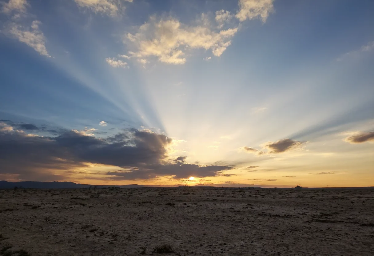 s2ms8ng, Vast barren landscape with cracked dry ground, dramatic sunset with golden light streaming through dark clouds, sunbeams radiating outward creating a striking pattern, soft pastel hues transitioning from yellow to blue in the sky, scattered wispy clouds enhancing the visual depth, silhouetted horizon with faint mountain range, tranquil and expansive atmosphere