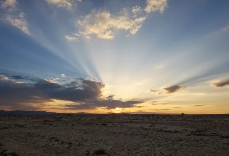 s2ms8ng, Vast barren landscape with cracked dry ground, dramatic sunset with golden light streaming through dark clouds, sunbeams radiating outward creating a striking pattern, soft pastel hues transitioning from yellow to blue in the sky, scattered wispy clouds enhancing the visual depth, silhouetted horizon with faint mountain range, tranquil and expansive atmosphere