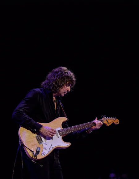 <lora:RICHIE_BLACMORE:1>The image is a photograph capturing a male musician performing on stage. He is centrally positioned, playing an electric guitar. The guitar is a light beige color with white accents, and he is holding it with both hands. The musician has shoulder-length, dark, wavy hair and is wearing a black, long-sleeved shirt. He is also adorned with several bracelets on his right wrist. His facial expression is focused and intense, indicative of deep concentration.
The background is dark, creating a stark contrast that highlights the musician. The lighting is focused on him, casting a soft glow that enhances the texture of his hair and the sheen on the guitar. The overall ambiance suggests an indoor concert setting, possibly a nightclub or small venue, given the intimate lighting and stage setup.
The photograph is a high-resolution, full-color image with sharp details, capturing the textures of the guitar and the musician's clothing. The image emphasizes the musician's connection with his instrument, showcasing the dynamic interplay between performer and instrument. The photograph captures a moment of artistic expression and musical engagement.