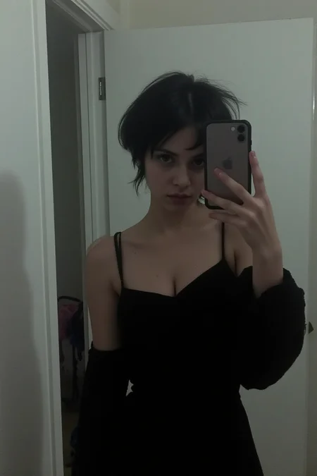 A 23-year-old goth petite woman stands in front of a foggy mirror, her short black hair framing her pale face. In a black bare shoulders dress, she snaps a moody selfie on the bathroom mirror, dirty mirror, her eyes downcast but still rebellious