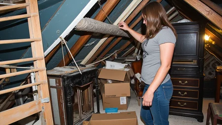 early 2010s snapshot photo captured with a phone and uploaded to facebook, A woman with shoulder-length brown hair, dressed in a t-shirt and jeans, navigates a cramped, dusty attic. Wooden beams, old furniture, and cardboard boxes surround her, emphasizing the confined space.
