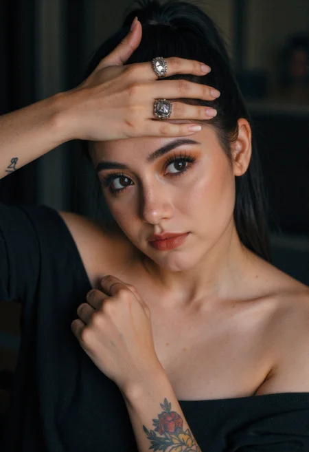 A woman with dark hair pulled back in a ponytail is looking to the left, with her right hand raised and her fingers near her forehead, wearing two large silver rings on her right hand. She has dark eye makeup, including eyeliner and eyeshadow, and a small dot on her forehead between her eyebrows. A floral tattoo is visible on her right forearm. She is wearing a dark top that is off the shoulders. The background is dark and out of focus.